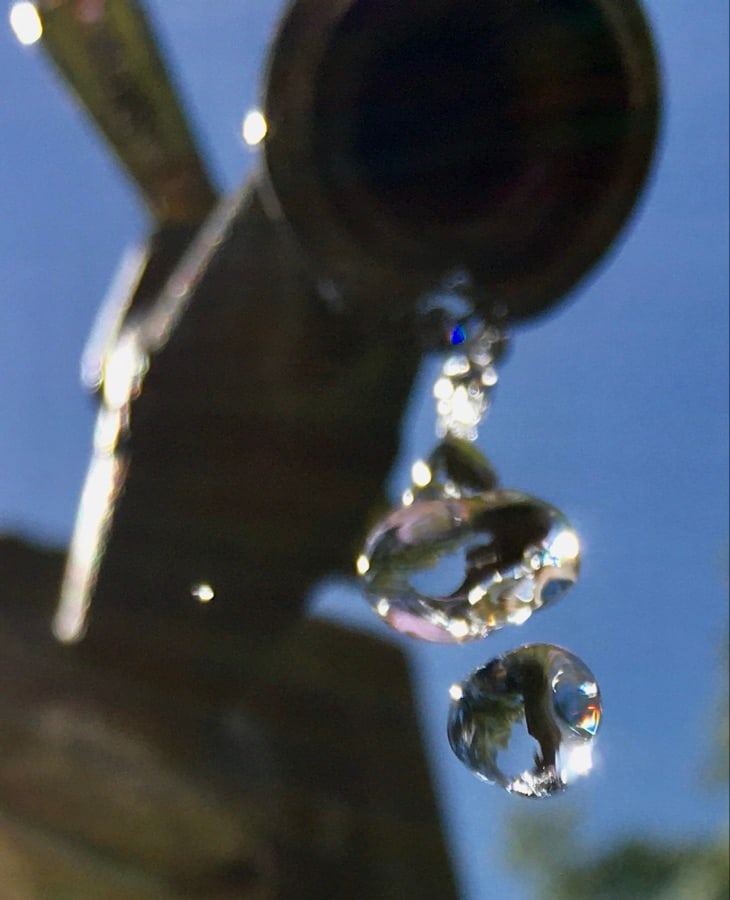 Close-up of two crystal-clear water drops falling from a metal spout with sky reflections