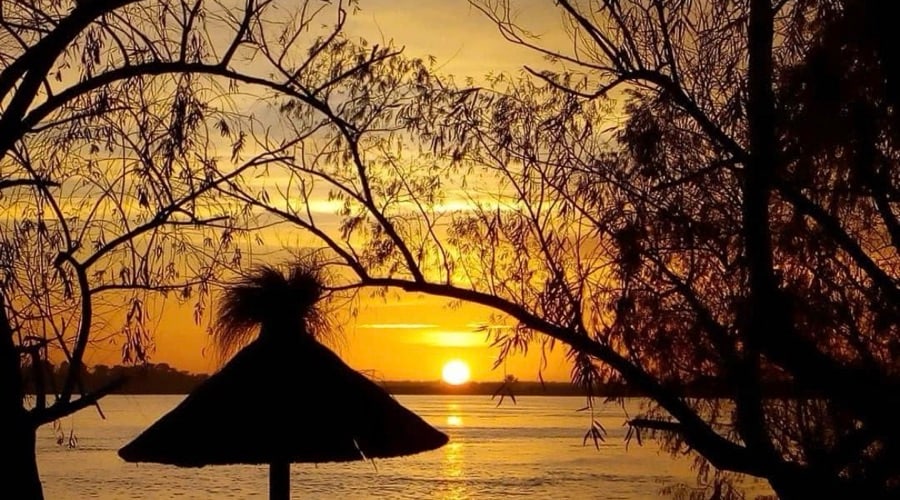 Atardecer sobre el río con siluetas de árboles y una estructura de techo de paja en primer plano