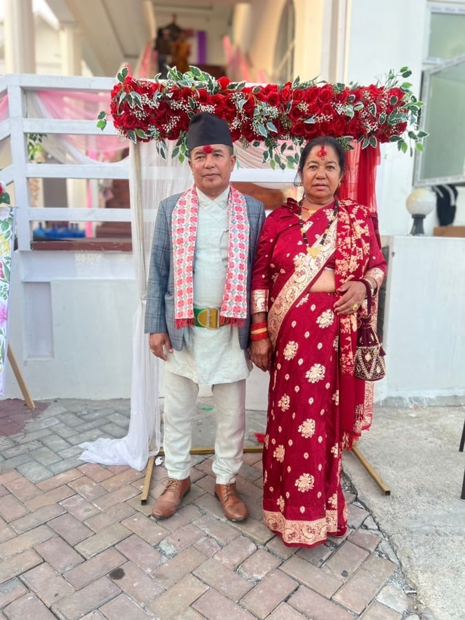 Formal portrait of a couple beneath a red floral arch, wearing traditional Nepali attire for a special occasion.