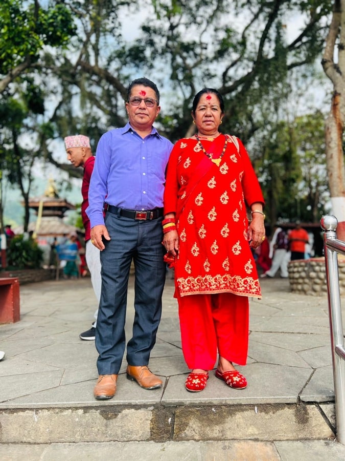 A couple standing outdoors in Kathmandu District, with the woman in bright red traditional dress and the man in a blue shirt and formal trousers.